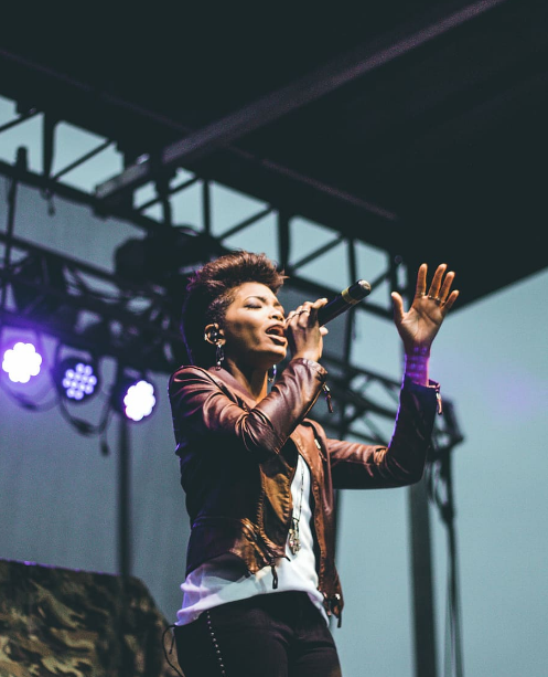 a black woman in a leather jacket singing live on stage in the open air